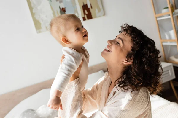 Femme joyeuse et bouclée en tenue de salon tenant dans les bras bébé fille en barboteuse — Photo de stock