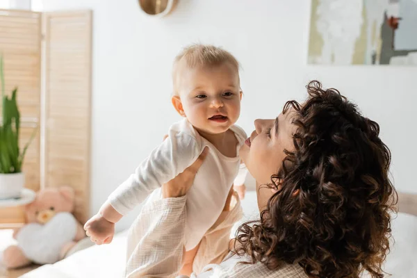 Happy and curly woman in loungewear holding in arms baby in romper — Stock Photo
