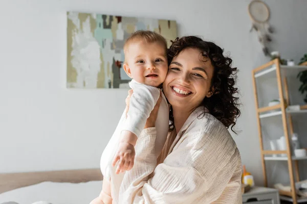 Happy and curly woman in loungewear holding in arms baby girl in romper — Stock Photo