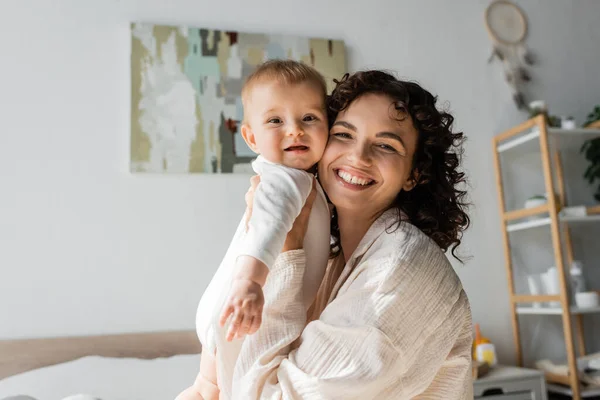 Glückliche und lockige Frau in Loungewear, die ein kleines Mädchen im Strampler im Arm hält — Stockfoto