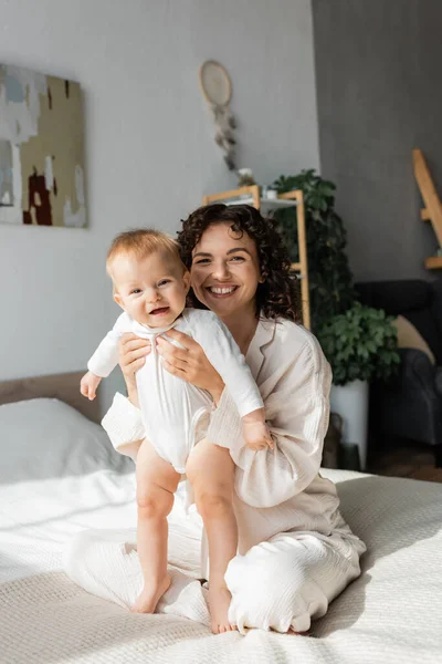 Happy and curly mother holding baby in romper and sitting on bed — Stock Photo