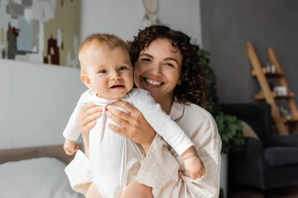Cheerful and curly mother holding daughter in romper in bedroom — Stock Photo