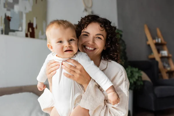 Joyeuse et frisée mère tenant fille dans la barboteuse dans la chambre — Photo de stock