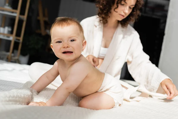Infant baby in romper sitting on bed near blurred and curly mother — Stock Photo