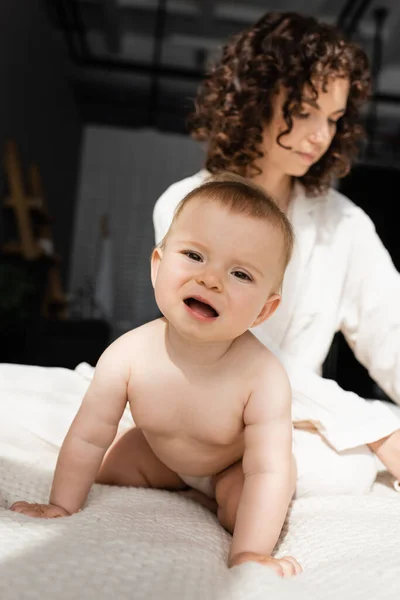Infant child in romper sitting on bed near blurred and curly mother — Stock Photo