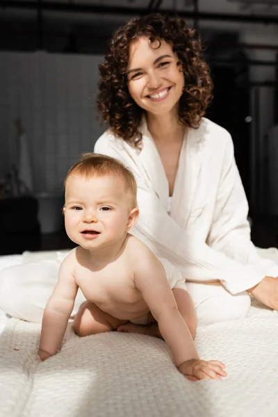 Infant child in romper sitting on bed near happy and curly mother — Stock Photo
