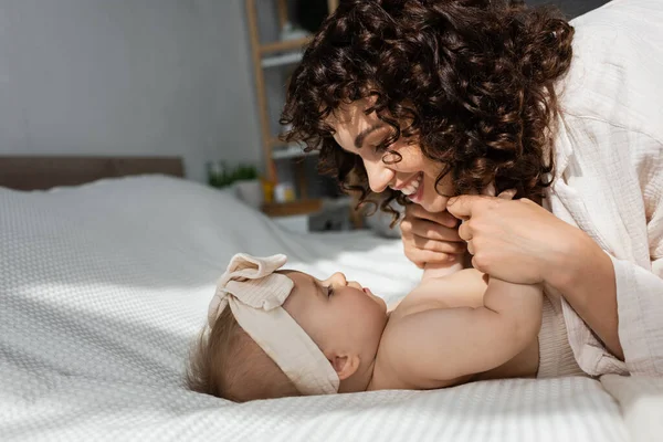 Visão lateral da mãe alegre com cabelo encaracolado segurando as mãos da menina na cabeça — Fotografia de Stock