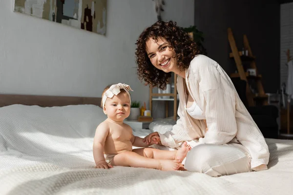Cheerful mother with curly hair sitting on bed with baby daughter in headband — Stock Photo