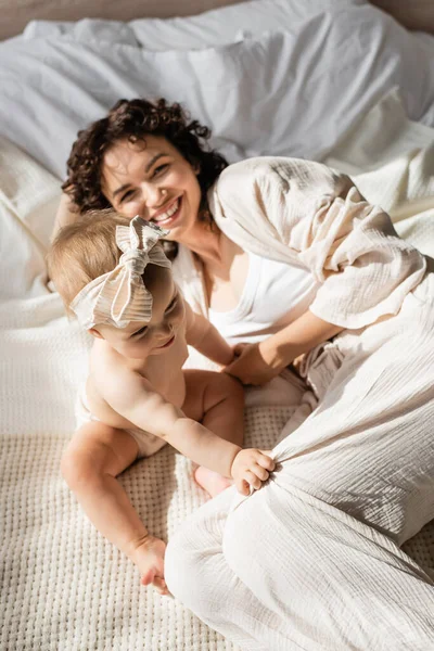 Vista ad alto angolo di madre allegra con capelli ricci sdraiati sul letto con bambina in fascia — Foto stock