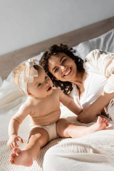 Fröhliche Mutter mit lockigem Haar liegt auf dem Bett neben süßer Baby-Tochter im Stirnband — Stockfoto