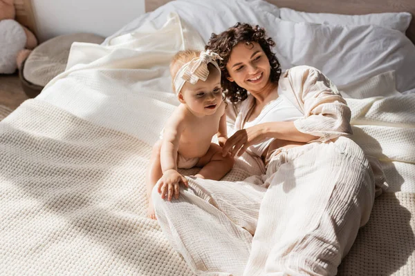 Feliz madre con el pelo rizado acostado en la cama cerca de linda hija lactante en diadema - foto de stock