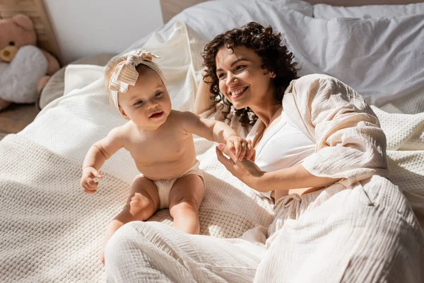 Madre feliz con el pelo rizado acostado en la cama y mirando a la hija lactante en diadema - foto de stock