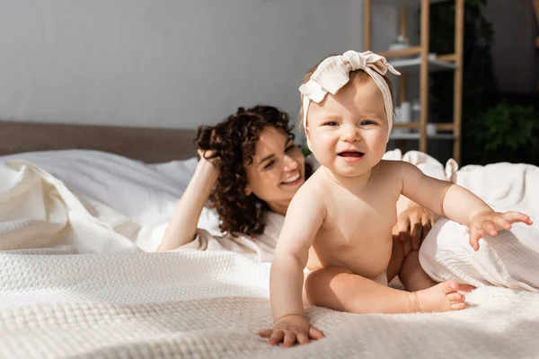 Маленька дівчинка в пов'язці сидить на ліжку біля щасливої матері з кучерявим волоссям — стокове фото