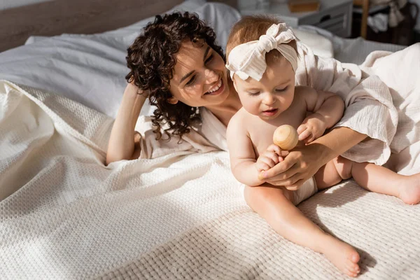 Donna positiva con capelli ricci sdraiata sul letto con figlia neonata in fascia che tiene in mano un giocattolo di legno — Foto stock