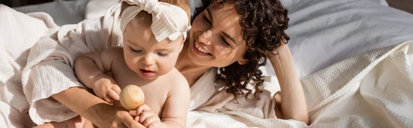 Happy woman lying on bed near infant daughter in headband with wooden toy, banner — Stock Photo