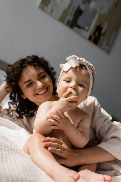 Радостная женщина с вьющимися волосами, лежащими на кровати и смотрящая на новорожденную дочь в повязке на голове с луком — стоковое фото