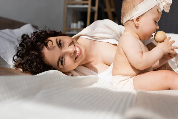 Happy mother lying on bed and looking at camera near baby girl with wooden rattle — Stock Photo