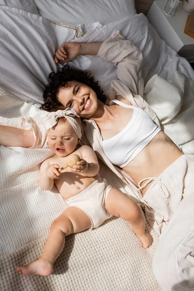 Ansicht von oben: Glückliche Mutter liegt mit Baby im Bett und hält Holzrassel — Stockfoto