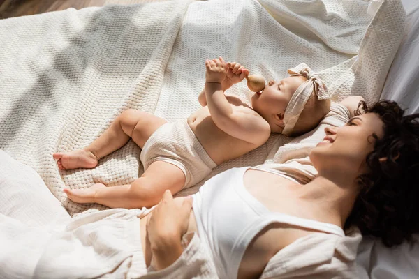 Вид сверху на счастливую мать, лежащую на кровати и смотрящую на девочку в головном уборе с бантом, держащим деревянный гремел — стоковое фото