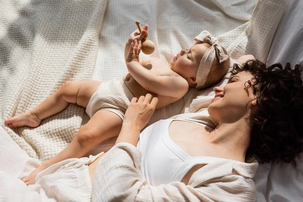 Draufsicht der fröhlichen Mutter, die auf dem Bett liegt und das kleine Mädchen im Stirnband mit Schleife ansieht, die eine Holzrassel hält — Stockfoto