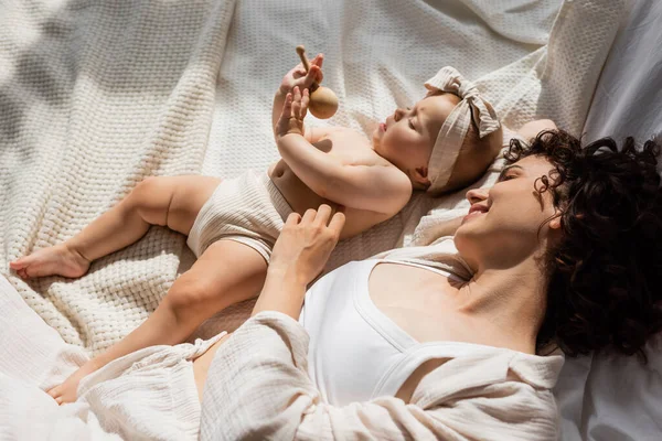 Top view of cheerful mother lying on bed and looking at baby girl in headband with bow holding wooden rattle — Stock Photo