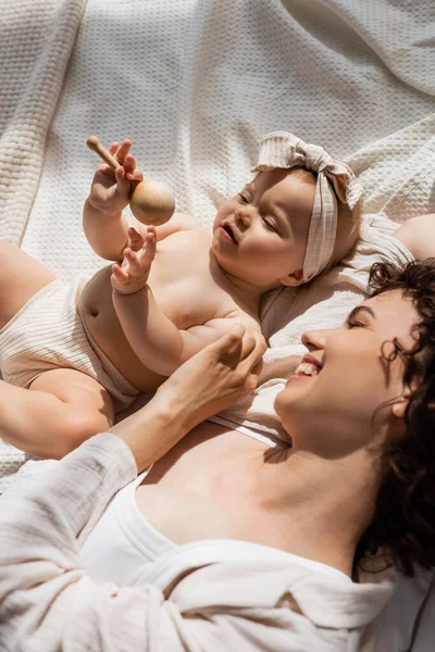 Top view of cheerful mother lying on bed near baby girl in headband holding wooden rattle — Stock Photo
