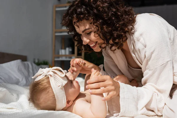 Glückliche und lockige Mutter hält die Hände eines kleinen Mädchens mit Stirnband, das zu Hause auf dem Bett liegt — Stockfoto
