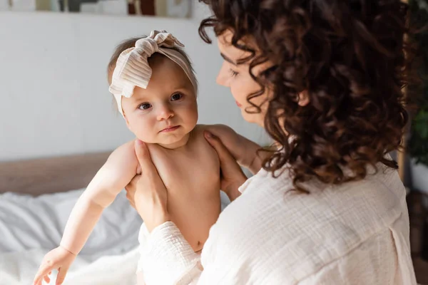 Curly mother holding in arms baby girl in headband with bow — Stock Photo