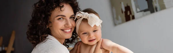 Cheerful and curly mother holding in arms baby girl in headband with bow, banner — Stock Photo