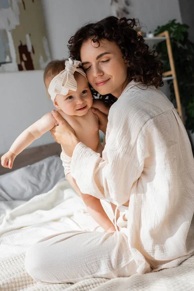 Curly mother with closed eyes holding in arms happy baby girl in headband with bow — Stock Photo