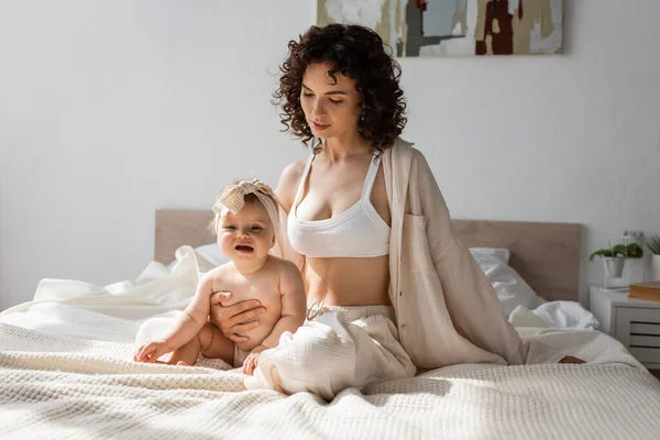 Mère bouclée en vêtements de salon avec le haut de la culture assis sur le lit avec la fille en bas âge dans le bandeau — Photo de stock