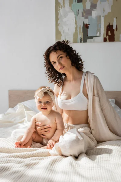 Кучерява жінка в лаунж-одязі з верхньою частиною врожаю сидить на ліжку з дочкою-немовлям в пов'язці — стокове фото
