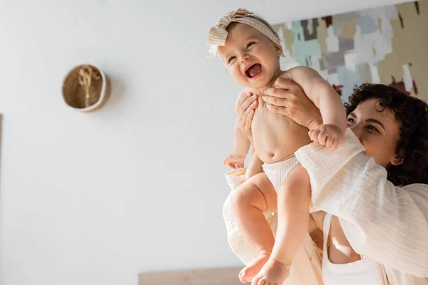 Curly woman holding happy infant daughter in headband above head in bedroom — Stock Photo