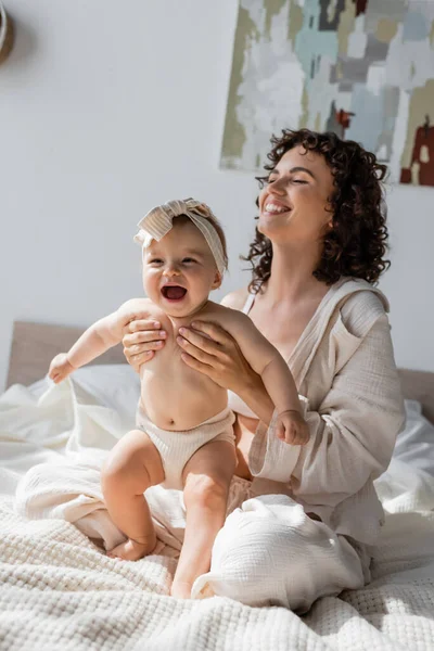 Mãe alegre em loungewear sentado na cama com a filha infantil feliz na cabeça — Fotografia de Stock