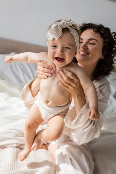 Alegre madre en ropa de salón sentada en la cama y sosteniendo feliz bebé hija en diadema - foto de stock
