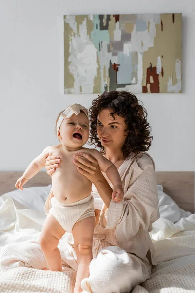 Curly mother in loungewear holding cute infant daughter in headband — Stock Photo