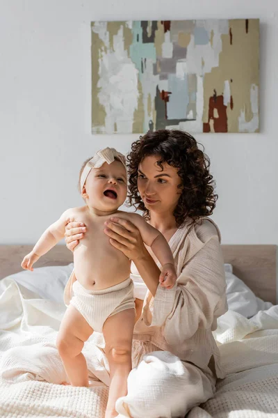 Mère bouclée en vêtements de salon tenant fille bébé mignon dans le bandeau — Photo de stock