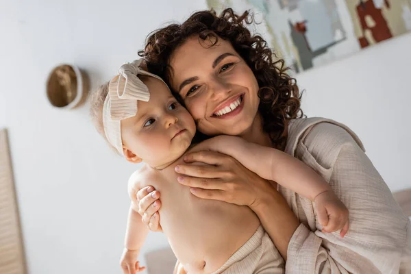 Madre rizada en ropa de salón sonriendo mientras abraza a la linda hija bebé en diadema - foto de stock