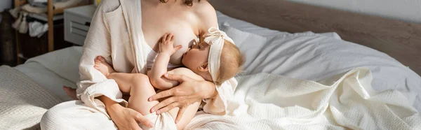 Madre in salottino seduta sul letto e che allatta figlia neonata in fascia con fiocco, stendardo — Foto stock