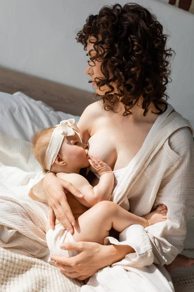 Mère bouclée en vêtements de salon assis sur le lit et l'allaitement bébé fille dans le bandeau avec arc — Photo de stock