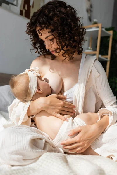 Caring mother in loungewear sitting on bed and breastfeeding infant daughter in headband with bow — Stock Photo