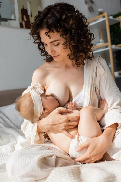 Mãe encaracolado em loungewear sentado na cama e amamentando filha infantil na cabeça — Fotografia de Stock