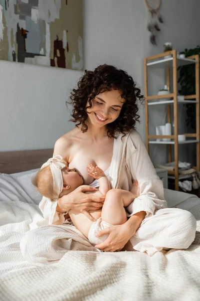 Mère heureuse en vêtements de salon assis sur le lit et l'allaitement bébé fille dans le bandeau avec arc — Photo de stock