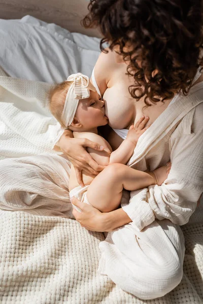 Високий кут зору кучерявої матері в одязі для відпочинку, що сидить на ліжку і годує немовлят дочка в пов'язці з бантом — стокове фото