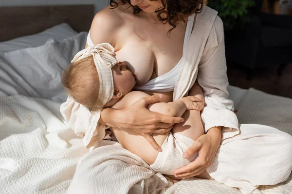 Curly woman in loungewear sitting on bed and breastfeeding infant daughter — Stock Photo