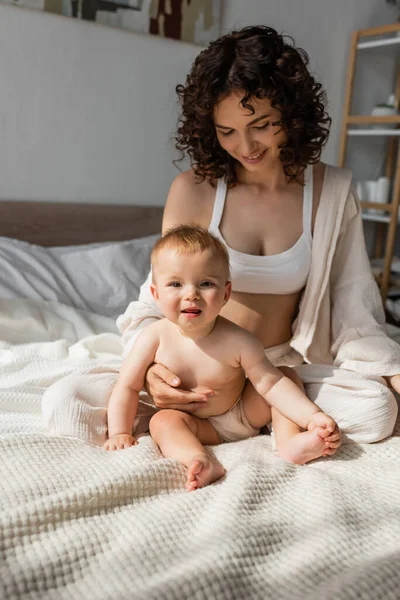Щаслива мати в лаунж-одязі з верхньою частиною культури, що сидить поруч з дочкою-немовлям у спальні — стокове фото