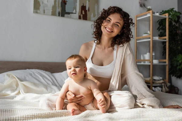 Fröhliche Mutter in Loungewear mit bauchfreiem Oberteil, die neben ihrer kleinen Tochter im Schlafzimmer sitzt — Stockfoto