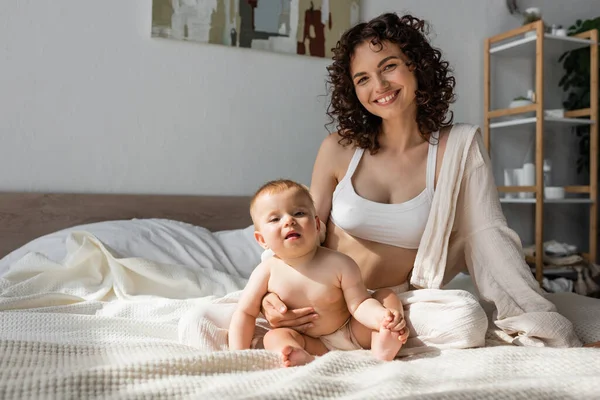 Позитивна мати в лаунж-одязі з верхнім посівом, що сидить поруч з дочкою-немовлям у спальні — стокове фото