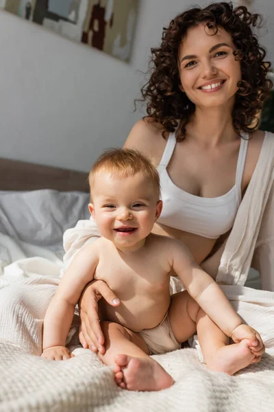 Радісна жінка в лаунж-одязі з верхнім посівом, що сидить поруч з дочкою-немовлям у спальні — стокове фото