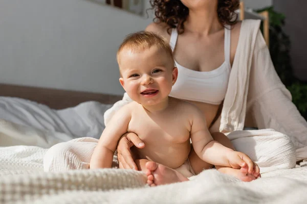 Madre in loungewear con crop top seduta vicino alla figlia infante felice in camera da letto — Foto stock